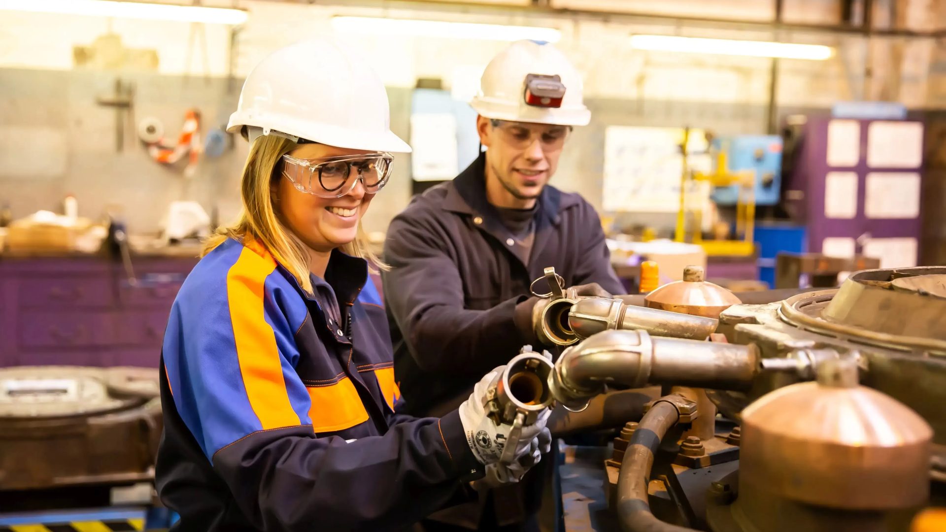 Zwei Ingeneure mit Schutzausrüstung arbeiten gemeinsam an Maschinenwartung in Industriewerkstatt.