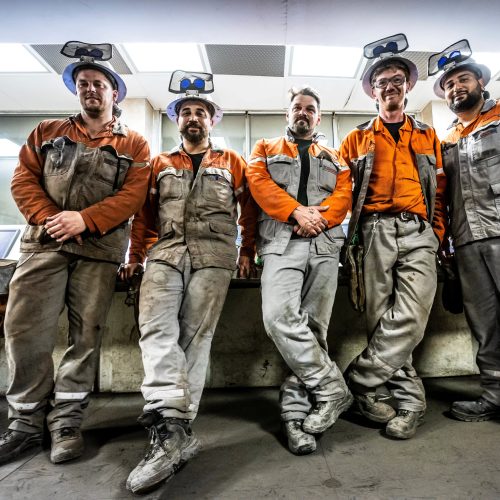 Team von Facharbeitern in Schutzkleidung und mit Helmen im Stahlwerk, vor Kontrollraum stehend.